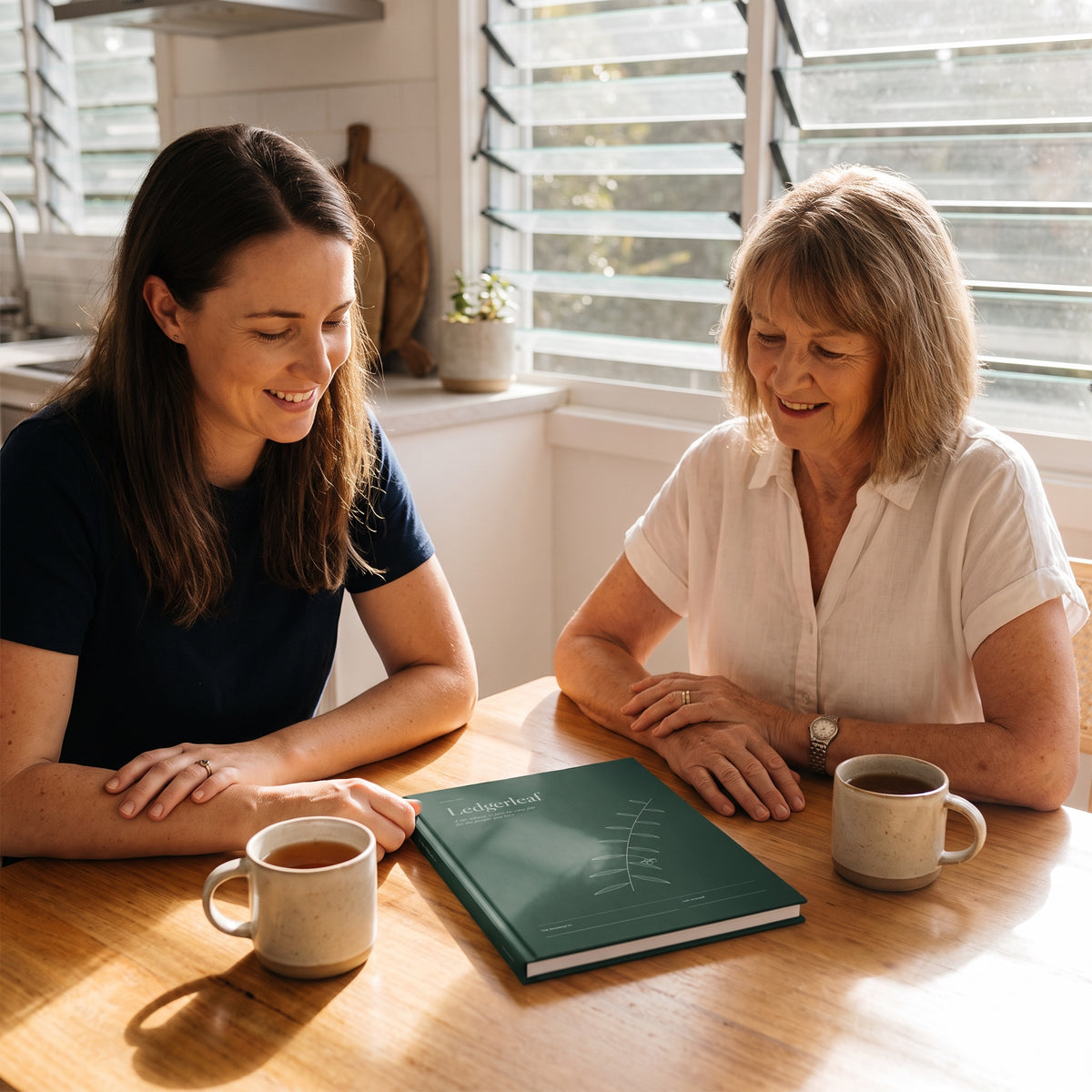 A mother and adult daughter at a sunny kitchen table with the Ledgerleaf life admin file between them